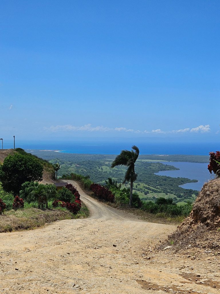 View from Montaña Redonda looking toward Miches, with a winding rural road, lush countryside, and glimpses of Laguna Redonda and Playa Esmeralda in the distance